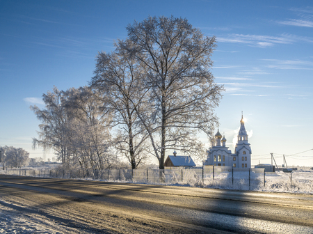 Orthodox white church with golden cupola and bell-tower next to the country road in sunny winter dayの写真素材