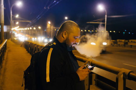 Vaping by electronic cigarette fat man using smart-phone on the bridge at night city.の写真素材
