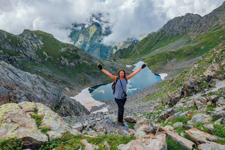 Joyful successful man tourist with raised arms on rock at mountains near cold mountain lake