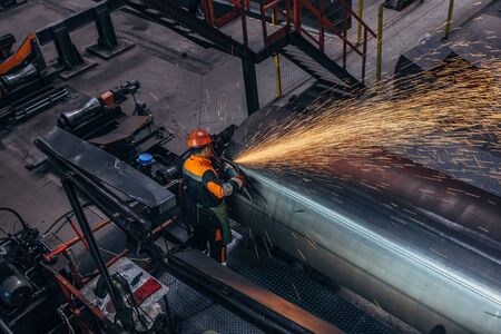 Worker cleans welded seam on steel pipe using grinding machine in metalwork workshop.の写真素材