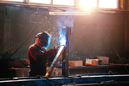 Factory worker in protective mask welding iron parts.