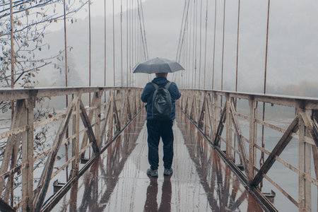 Man with umbrella walking on hanging bridge in rainy weather