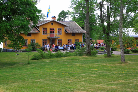 View of a yellow village house with swedish flag on the roof. Many guests coming for an outdoor party on a summer day. Beautiful swedish nature.のeditorial素材