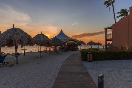 Beautiful view of coast line hotel area on sunset. View of tourists in line for drinks in outdoor bar. White sand coast and palm plants on sunset background. 17.17.2019. Oranjestad. Aruba.のeditorial素材