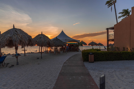Beautiful view of coast line hotel area on sunset. View of tourists in line for drinks in outdoor bar. White sand coast and palm plants on sunset background. Oranjestad. Aruba.のeditorial素材