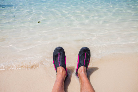 Close up view of rolling waves on men's feet standing on white sand beach in swimming shoes.