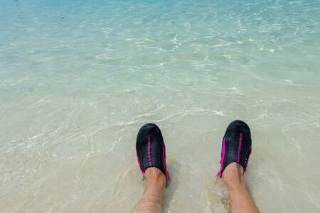 Close up view of rolling waves on men's feet standing on white sand beach in swimming shoes. Curacao island. Gorgeous nature landscape backgrounds. Caribbean.の写真素材