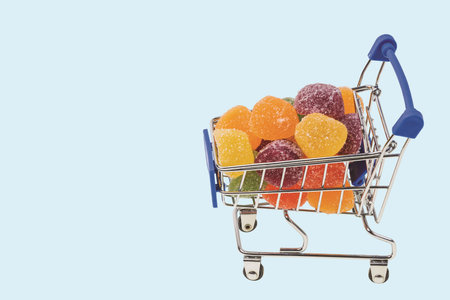 Close up view of colorful marmalade in shopping cart on blue background isolated. Unhealthy food...