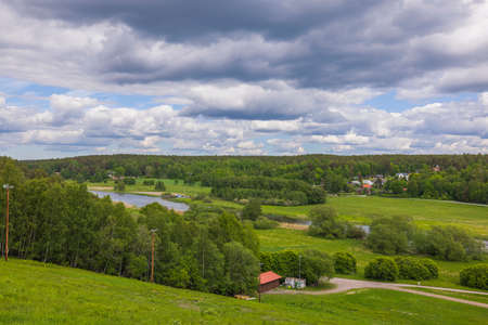 Amazing green summer nature landscape view. Fiew buildings on green fields between green forest trees under blue sky with puffy white clouds. Sweden.の写真素材