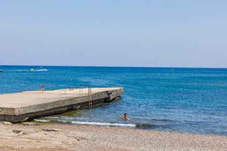 View of little girl sitting in water on coastline on hot summer day. Mediterranean. Greece. Rhodes. 08.25.2022.のeditorial素材