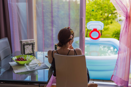 Young girl with hair decoration sitting in front of open window and looking outside. Inflatable swimming pool outside house. Sweden.の写真素材