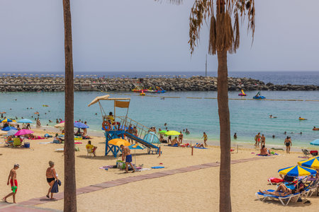 Sandy shore of Atlantic Ocean in Gran Canaria with tourists relaxing on equipped beach, featuring umbrellas and loungers, along with lifeguard station.のeditorial素材