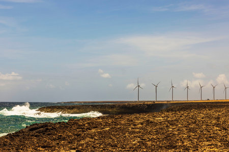 Wind turbines along rocky coastline with ocean waves and blue sky in background. Aruba.の写真素材