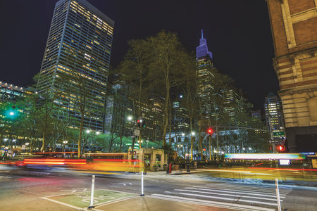 Nighttime view of a busy city street with light trails from cars against skyscrapers and trees under...