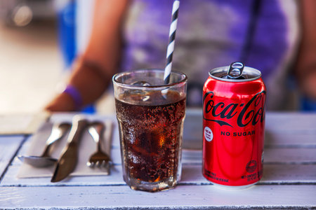 Glass of cola with ice and striped straw alongside can of Coca-Cola No Sugar on table with utensils.のeditorial素材