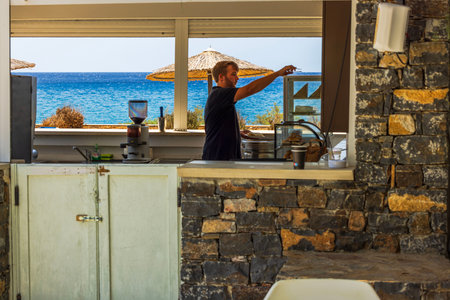 Man preparing food in seaside cafe with stone walls and view of Mediterranean sea in background. Crete. Greece.のeditorial素材