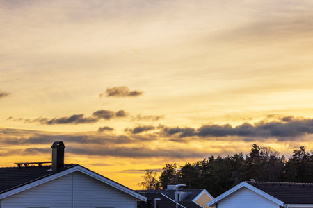 Silhouetted rooftops and tree line against golden sunset sky with scattered clouds.の写真素材