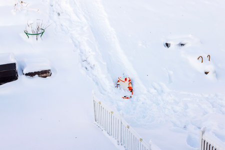 Bright snowy backyard with orange plastic sled left in deep snow near white picket fence during winter day.の写真素材