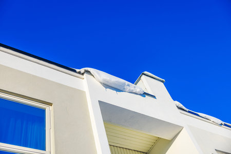 Snow over edge of white house roof under hanging clear blue sky on sunny winter day.