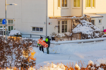 Children standing in snowy street holding colorful sled near residential house during bright sunny winter day.のeditorial素材
