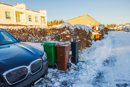 Snow-covered residential street with parked black car, green and brown garbage bins and hedges in winter day. Sweden.の写真素材