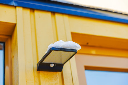 Solar outdoor LED light with motion sensor and snow resting on its surface mounted on yellow wooden wall villa.の写真素材