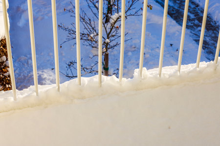 Snow-covered railing on villa balcony overlooking snowy tree in garden and pathway during bright winter day.の写真素材