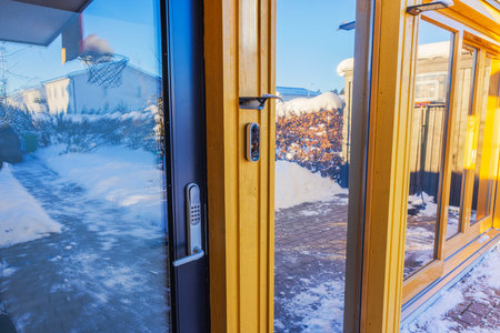 Modern glass door with digital lock and security camera on yellow wooden frame, reflecting snowy yard and basketball hoop. Sweden.の写真素材