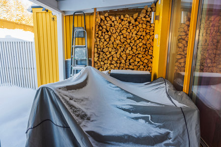 Snow-dusted patio with stacked firewood, covered outdoor grill and metal ladder against bright yellow wooden wall villa.の写真素材