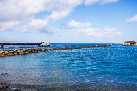 Pier extending into clear blue water Caribbean sea with rocky shoreline and thatched hut in Curacao against on cloudy sky background.の写真素材