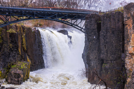 Great Falls waterfall in Paterson, New Jersey, cascading powerfully over rocky cliffs with an iron bridge above.の写真素材
