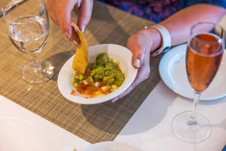 Hand holding tortilla chip dipping into fresh guacamole and salsa in white bowl surrounded by wine and water glasses. Mexico.の写真素材