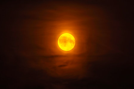 Bright full moon glowing in dark night sky surrounded by thin clouds with an orange hue.の写真素材