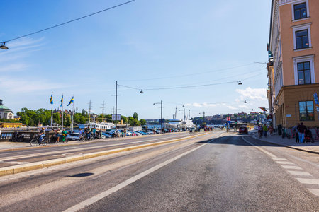 Street in central Stockholm with Nordstrom river, Swedish flags, pedestrians, bicycles, and ships visible along the waterfront. Sweden. Stockholm.のeditorial素材