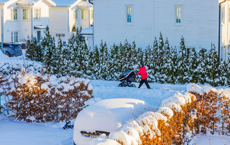 Woman pushing baby stroller on snowy street near white houses and hedges during winter day.のeditorial素材