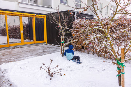 Child in winter outfit sitting on snow-covered ground garden near leafless apples trees by modern villa with panoramic reflective windows. Sweden.の写真素材