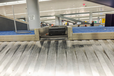 Black suitcase emerging on baggage carousel at modern airport terminal with passengers waiting in background. USA. New York.のeditorial素材