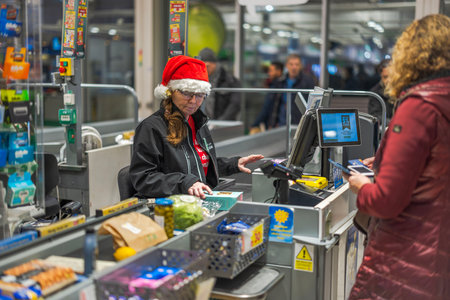 Cashier wearing Santa hat scanning groceries at supermarket checkout counter while customer waits with purchases. Sweden. Uppsala. 12.23.2024.のeditorial素材