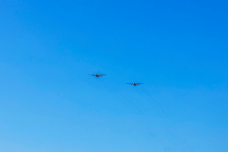 Two military transport planes flying parallel in clear blue sky leaving faint smoke trails behind.の写真素材