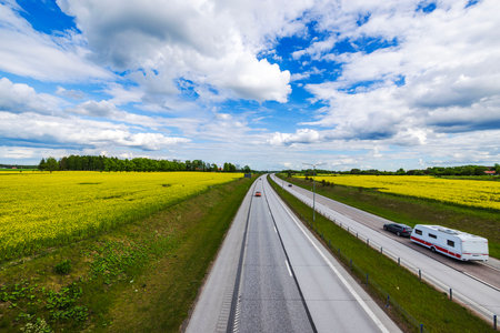 Straight highway cutting through yellow rapeseed fields under partly cloudy sky with cars and caravan driving.の写真素材