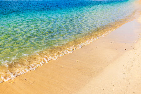 Crystal-clear turquoise water gently washing onto golden sandy beach under bright sunlight creating a tranquil scene.の写真素材