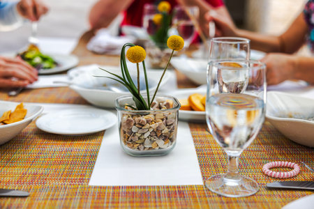 Dining table setting with yellow flowers centerpiece, water glasses, plates and colorful mat during meal. Mexico.の写真素材