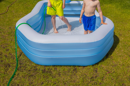 Two boys standing in an inflatable pool on green grass with garden hose on sunny summer day.の写真素材