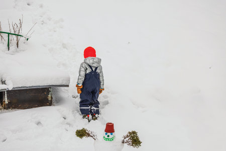 Child in silver jacket and red hat walking through snowy backyard near small snowman during winter day. Sweden.の写真素材