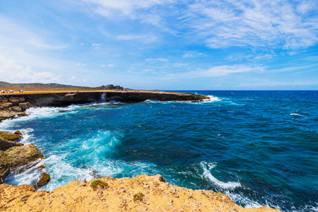 Rocky coastline of Aruba with turquoise waves crashing against cliffs, ruins of Bushiribana Gold Mill plant visible on horizon.の写真素材