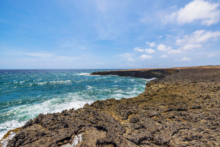 Waves crashing against rugged volcanic coastline of Arikok National Park in Aruba, with wind turbines standing along horizon under bright blue sky.の写真素材