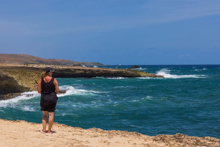 Woman standing on rocky shore of Arikok National Park in Aruba, gazing at waves crashing against rugged coastline of Caribbean Sea.の写真素材