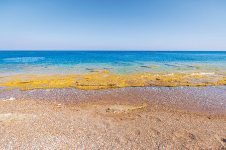 Clear turquoise water of Aegean Sea washing over rocky shore with small pebbles under bright sunlight.の写真素材