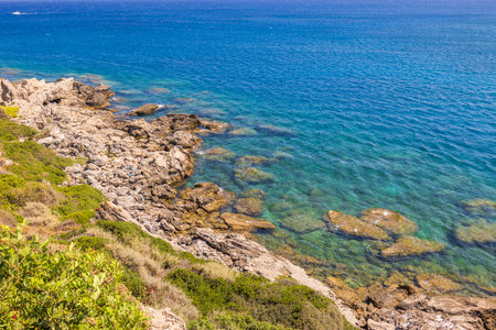 Rocky coastline with clear turquoise water of the Aegean Sea and green vegetation under bright sunlight.の写真素材