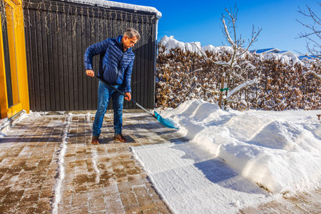 Close up view of man clearing snow from brick patio in front of villa entrance on sunny winter day. Sweden.の写真素材